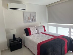 A bedroom with a bed, a nightstand, a lamp, an air conditioner, a window with a blind, and a cityscape painting on the wall. - Apartamento (Santa Marta)