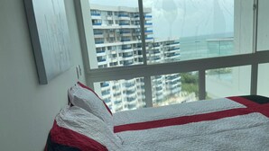 Un dormitorio con cama, ventana que ofrece vista a un edificio y un cuadro en la pared.