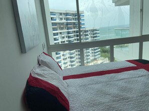 A bedroom with a bed, a window with a view of a building, and a wall with a picture.