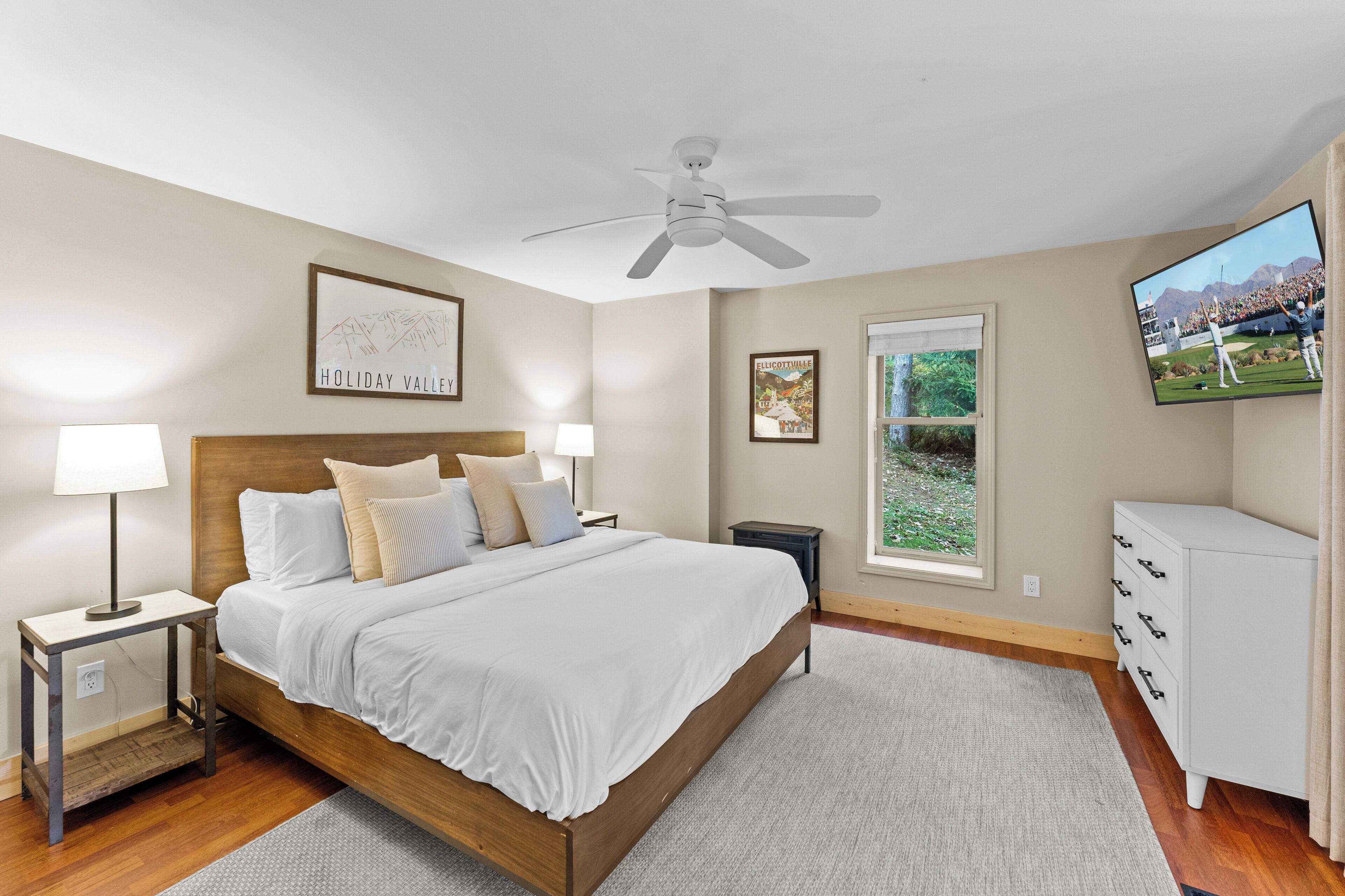A bedroom with a wooden bed, two bedside tables with lamps, a ceiling fan, a TV, and a window with a view of greenery.