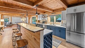 A spacious kitchen with wooden beams, a large island with stools, a stainless steel refrigerator, and a view of the outdoors. - Beautiful chalet on 5 acres - 2 min to Village!👌 Hot-Tub, Sauna, Fire Pit🔥 (Ellicottville)
