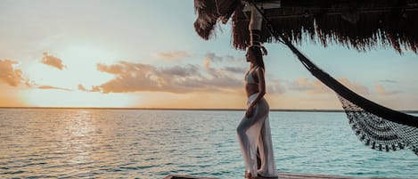 A wooden deck with a thatched roof overlooking a calm sea at sunset.