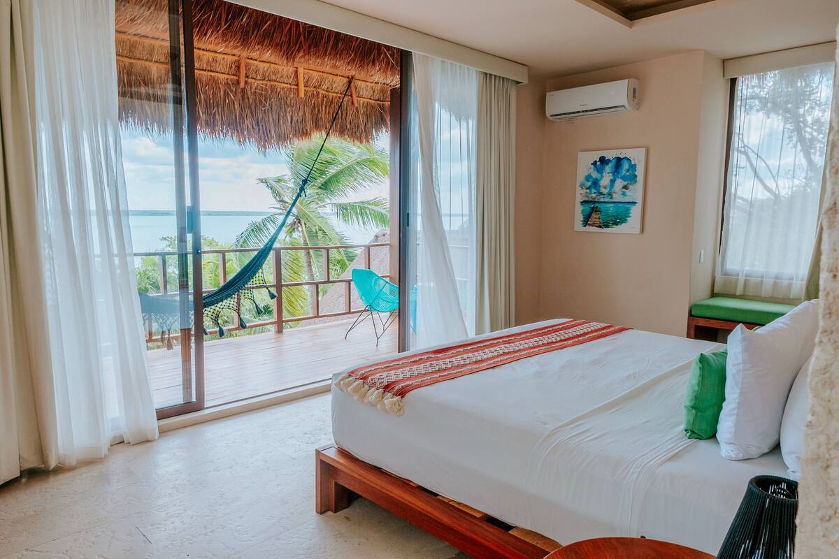 A bedroom with a large bed, a view of the ocean, a hammock, and a chair outside the window.