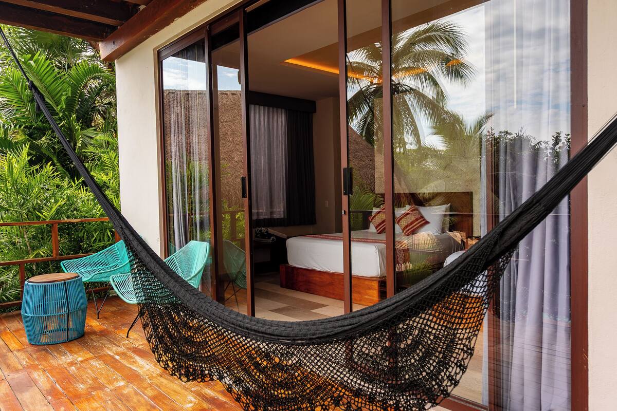 A hammock on a wooden deck with a sliding glass door leading to a bedroom with a bed and curtains.