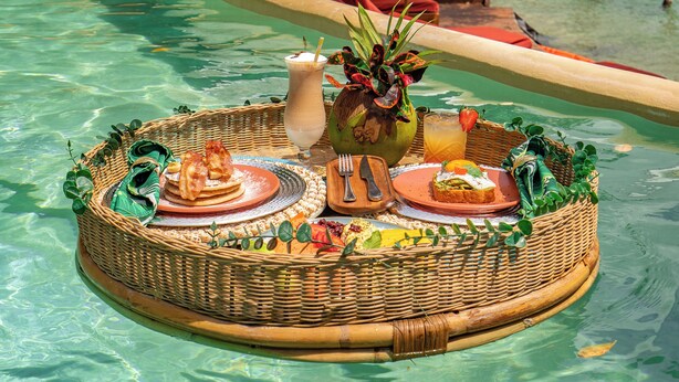A floating dining setup with a wicker table, plates, cutlery, and a drink in a tropical setting. - Khaban Bacalar Hotel Boutique (Bacalar)