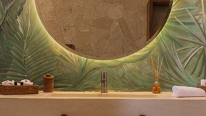 A bathroom with a large circular mirror, a sink, a basket of toiletries, and a bottle of reed diffuser. - Khaban Bacalar Hotel Boutique (Bacalar)