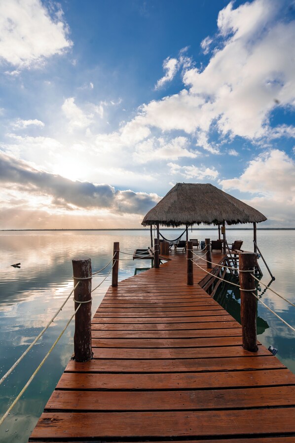 A wooden pier leading to a thatched-roof structure with seating, overlooking calm waters and a partly cloudy sky. - Khaban Bacalar Hotel Boutique (Bacalar)
