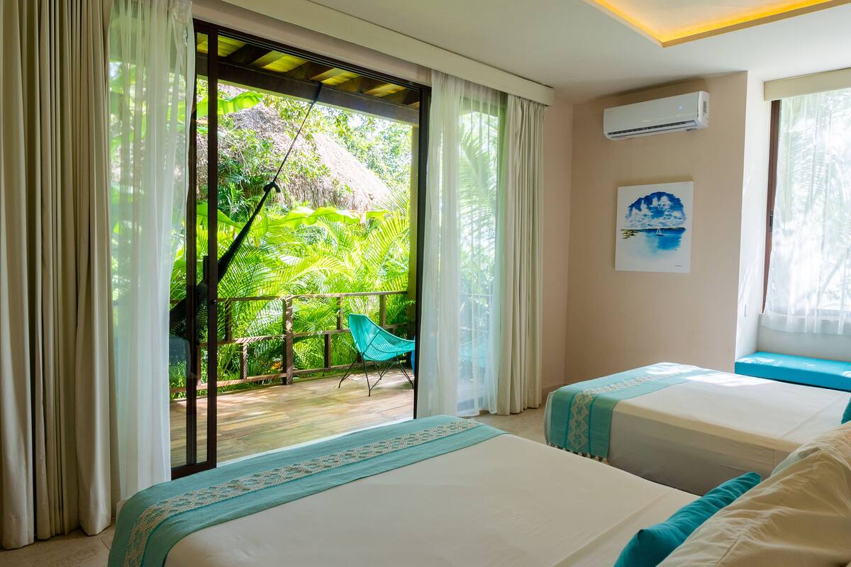 A hotel room with two beds, a balcony with a chair, and a view of lush greenery.