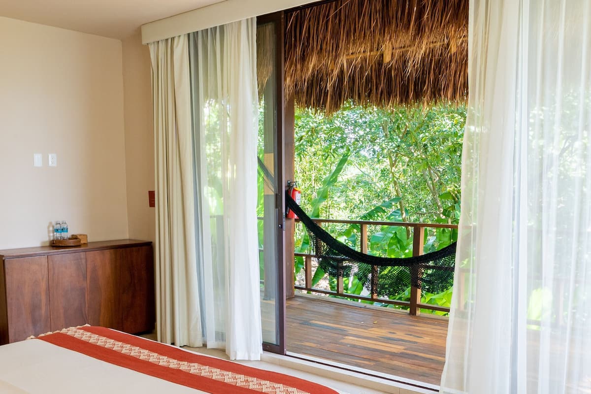 A bedroom with a bed, a wooden bench, a nightstand, a ceiling fan, and a view of a hammock and greenery outside.