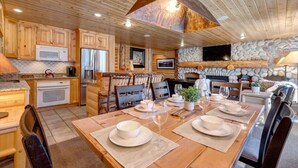 A wooden cabin interior with a dining area, kitchen, and stone fireplace.