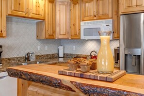 A kitchen with wooden cabinets, a granite countertop, a stainless steel refrigerator, a microwave, and a wooden cutting board with muffins and a pitcher of juice.