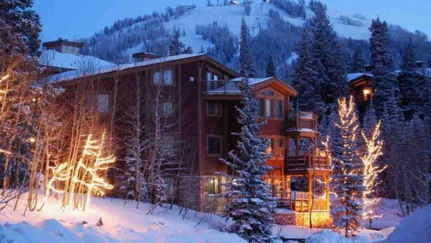 A lodge with a wooden exterior, multiple stories, and a balcony. It is surrounded by snow-covered trees and a mountain backdrop.