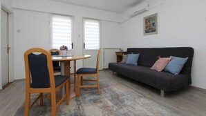 Ein kleines, helles Wohnzimmer mit einem grauen Sofa, zwei Holzstühlen, einem runden Tisch und einem gemusterten Teppich.