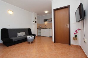 A living room with a sofa, a small round table, a door, a TV mounted on the wall, and a kitchen in the background.