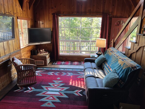 A cozy cabin interior with a wooden interior, a sofa, a TV, and a window with a view of greenery. - Cabin in Lake Arrowhead, walk to Lake and Village (Lake Arrowhead)