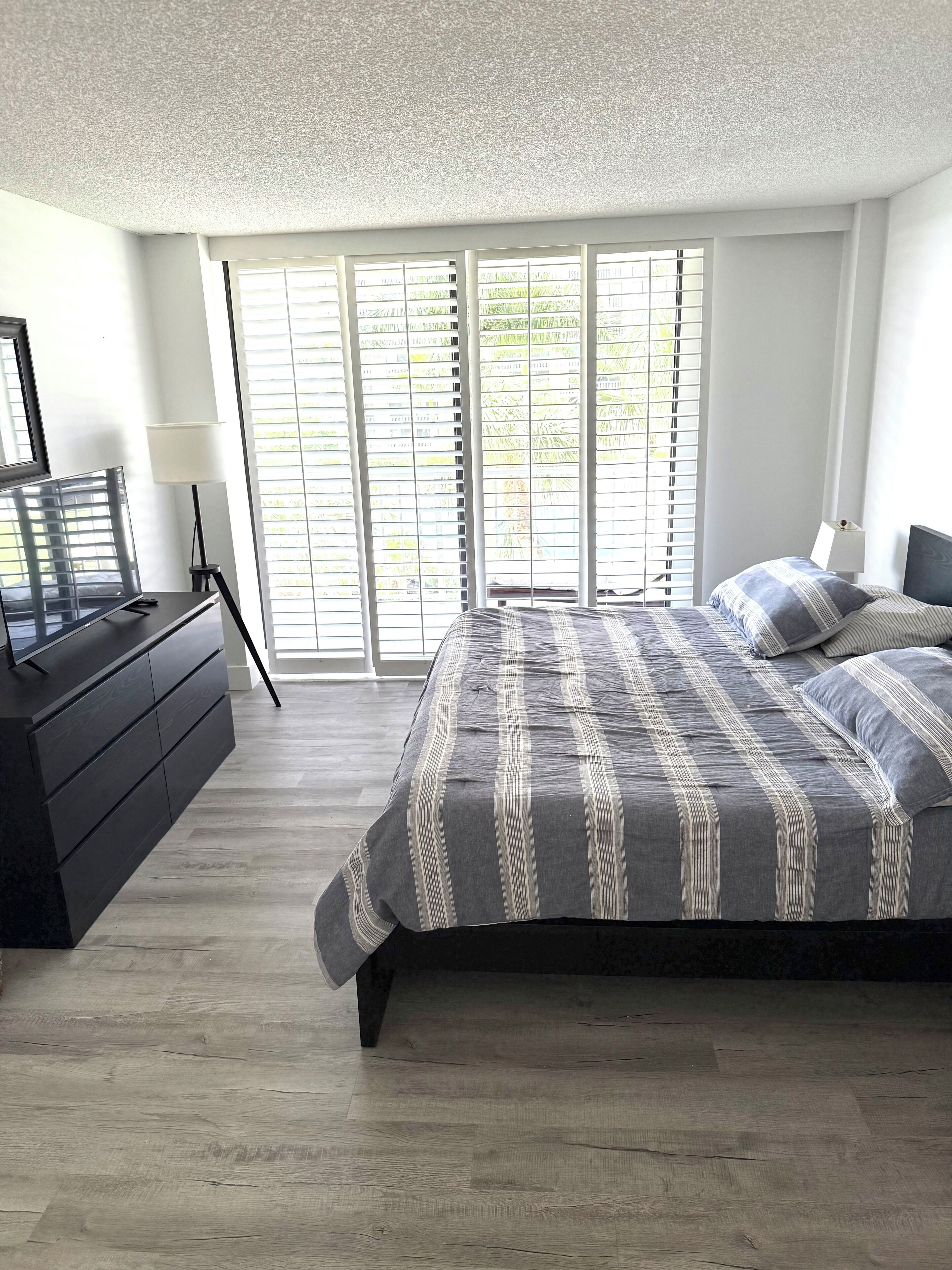 A bedroom with a large bed, a dresser, a TV, and a window with blinds.