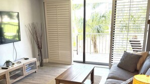 A living room with a sofa, a coffee table, a TV, and a sliding glass door leading to a balcony. - Beach Front Condo in Paradise Marco Island (Marco Island)