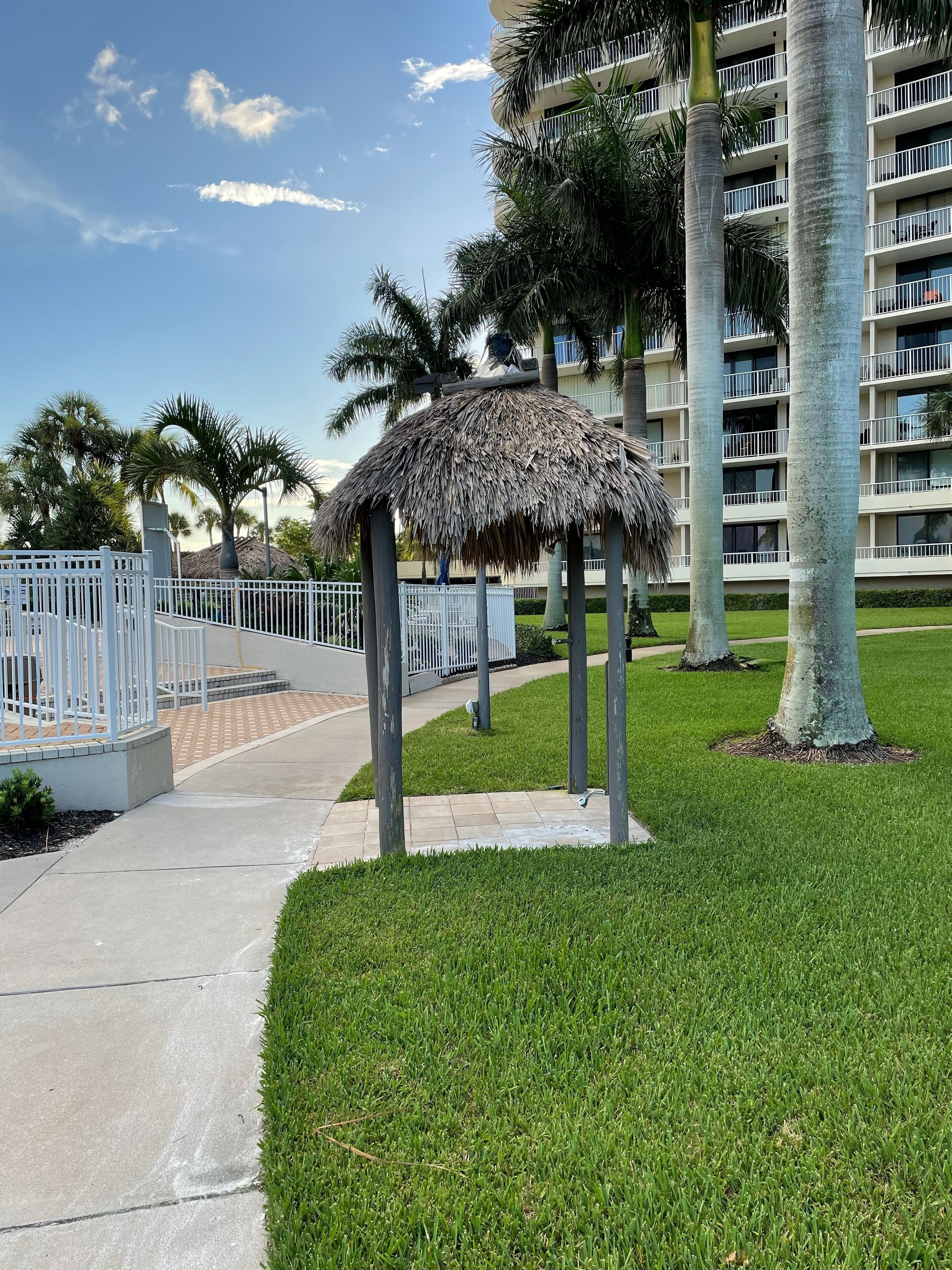 A thatched gazebo, palm trees, a white fence, and a multi-story building with balconies.