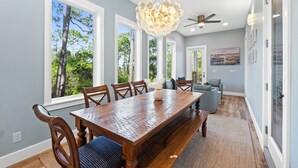 A dining area with a wooden table and chairs, large windows with a view of trees, a ceiling fan, and a chandelier.