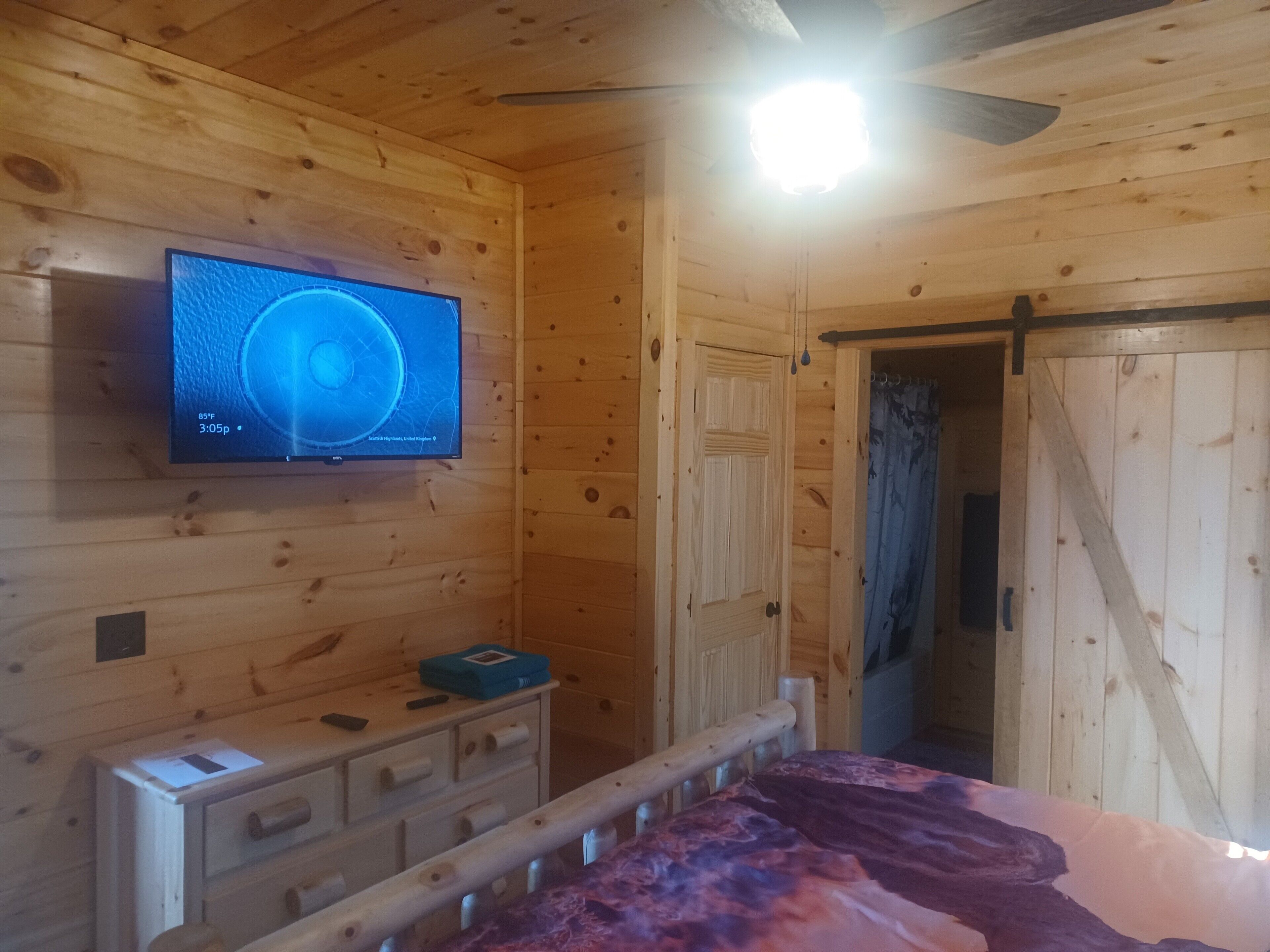 Une pièce de style cabane en bois, avec un téléviseur fixé au mur, un lit recouvert d’une courtepointe à motifs, une commode en bois et une porte donnant sur une autre pièce.