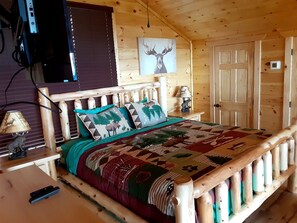 Una habitación de cabaña de madera con una cama, una lámpara, un cuadro de un alce y una puerta.