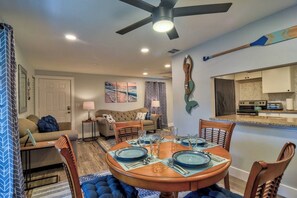 A dining area with a round table set for four, a ceiling fan, and a kitchen in the background.