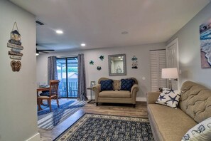 A living room with a sofa, a dining table with chairs, a patterned rug, and a view through a glass door.