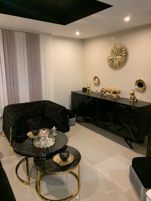A modern living room with a black sofa, a glass coffee table with gold legs, a black console table with decorative items, and a large gold wall art piece.