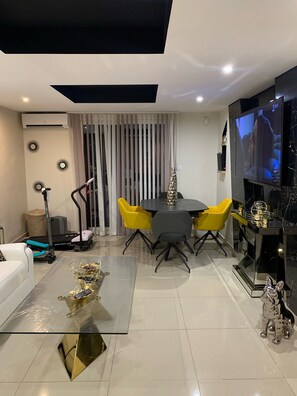 A modern living room with a dining area, a TV mounted on the wall, and a treadmill in the corner.