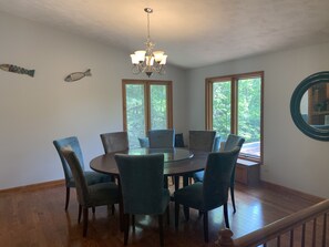 Un comedor con una mesa redonda, seis sillas, una araña, dos ventanas y un espejo en la pared.