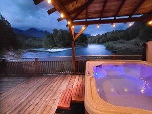 A hot tub on a wooden deck with a view of a river and mountains.