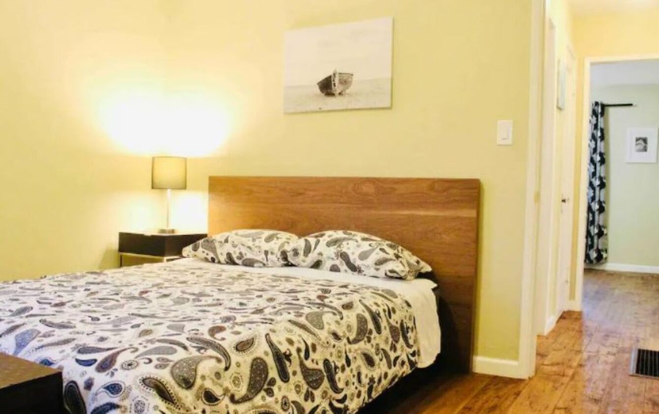 Un dormitorio con cabecera de madera, colcha estampada, lámpara de noche y un cuadro enmarcado en la pared.