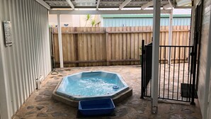 A hot tub in a covered outdoor area with stone flooring, a wooden fence, and a metal gate.