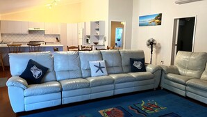 A modern living room with a grey leather sofa, a dining area with wooden chairs, and a kitchen in the background.