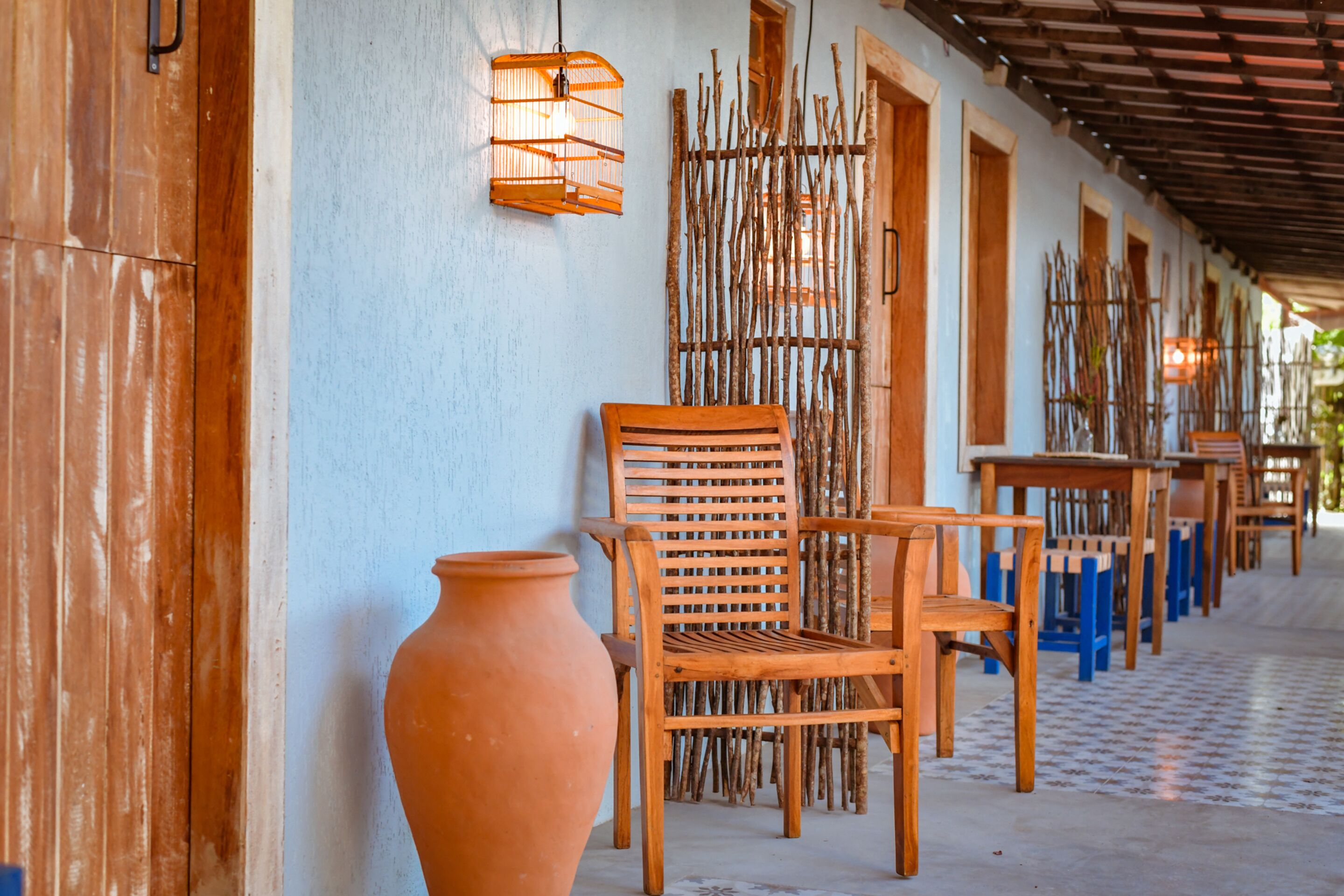Un pasillo con sillas y mesas de madera, una maceta de barro grande y un adorno de pared de bambú.