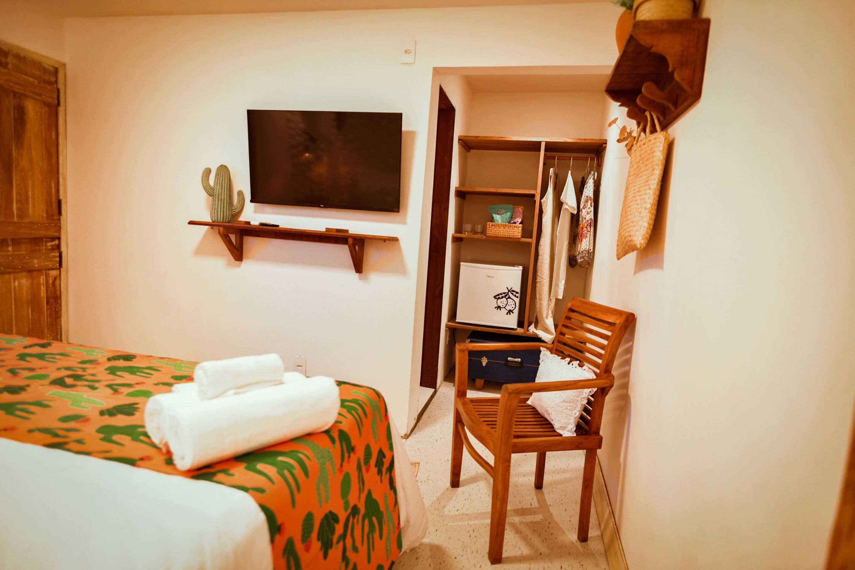 A hotel room with a bed, a wooden chair, a television, a wardrobe, and a cactus decoration.