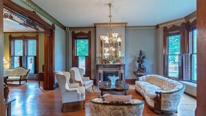 A spacious living room with a fireplace, two white armchairs, a patterned sofa, and a chandelier.
