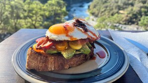 Un plato de desayuno con huevos, tocino, aguacate y tomates sobre tostadas, sobre una mesa con una vista panorámica de un río y colinas al fondo.