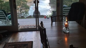 Un área de comedor con una mesa y sillas, una vela sobre la mesa, y vistas a un río y un bosque a través de puertas de cristal grandes.