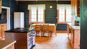 A kitchen with a stove, refrigerator, and wooden cabinets. There are two windows with lace curtains and a table with chairs.