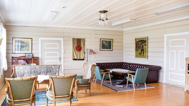 A cozy room with a wooden floor, a leather sofa, and several chairs. There are framed pictures on the walls and a ceiling light fixture.