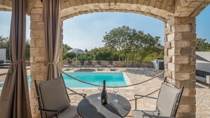 Ein Poolbereich mit einem Steinkunstwerk, zwei Stühlen, einem Tisch mit einer Flasche und Weingläsern sowie Blick auf den Pool und Bäume.
