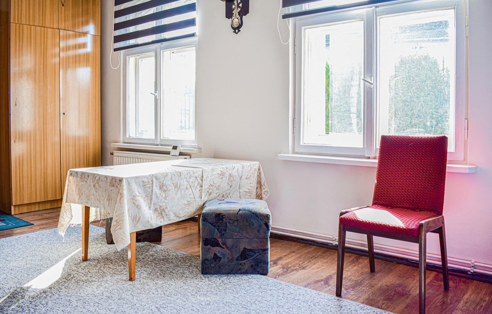 A room with a wooden wardrobe, a table with a floral tablecloth, a red chair, and a window with a view of trees.