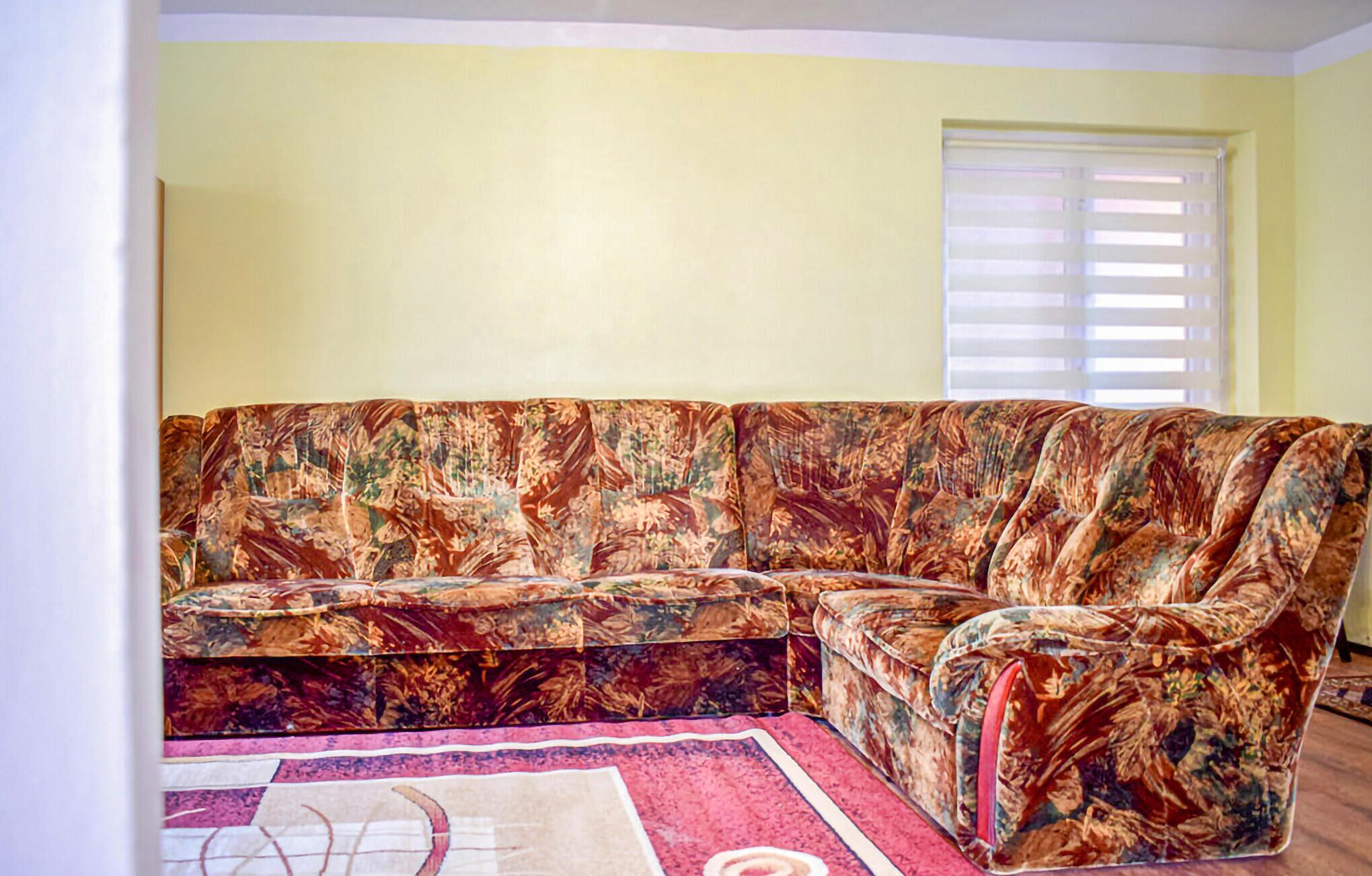 A sectional sofa with a floral pattern, a red and white rug, and a window with blinds.