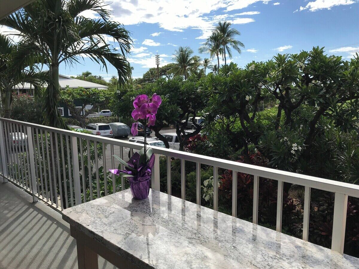 Un balcon avec une balustrade, un pot de fleurs contenant des fleurs violettes, et une vue sur des palmiers et des voitures garées.