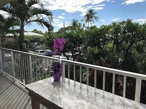 A balcony with a railing, a potted plant with purple flowers, and a view of palm trees and parked cars. - Puako Condo 205 ~ 3 BR ~ Renovated ~ Pool (Waimea)