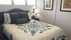 Ein Bett mit floralem Überwurf, ein Nachttisch mit Lampe sowie gerahmte Kunstwerke an der Wand.