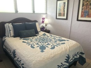 A bed with a floral-patterned bedspread, a nightstand with a lamp, and framed artwork on the wall. - Puako Condo 205 ~ 3 BR ~ Renovated ~ Pool (Waimea)