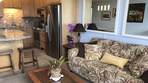 Ein Wohnzimmer mit einer Couch, einem Couchtisch mit einer Pflanze, einer Küchenzeile inklusive Kühlschrank und einem Essbereich mit Hockern.