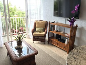 A living room with a glass-top coffee table, a wicker armchair, a wooden bookshelf, and a television mounted on the wall.